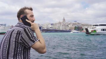 Mann reden auf das Telefon durch das Meer. jung Mann Sitzung auf das Bank gegen das Meer Gespräche auf das Telefon und Uhren das Meer. video