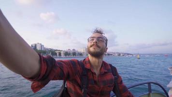 jovem homem levando selfie com telefone em balsa. jovem homem levando selfie com telefone contra mar e cidade visualizar. video