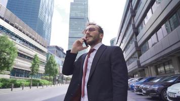 bärtig Mann Gehen entlang das Stadt Straße und sich unterhalten durch Telefon gut aussehend lächelnd bärtig Mann Gehen auf Stadt Straße, reden auf Zelle Telefon beim das gleich Zeit. video