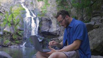 Freelance man outdoors. Young man at waterfall in nature working with laptop and looking around. video