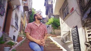 Happy young man is sitting on the street. A young man sits on the street with historic houses, looking around. video