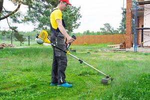 Male gardener mows the green grass of the lawn in the backyard at construction site with a gasoline mower. Trimmer for the care of a garden plot photo
