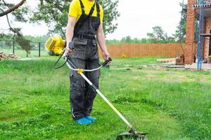 Male gardener mows the green grass of the lawn in the backyard at construction site with a gasoline mower. Trimmer for the care of a garden plot photo