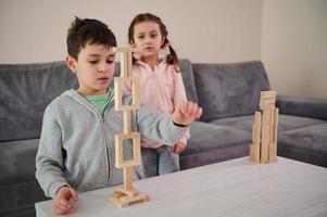 Focus on handsome school aged boy building tall structure of frames with wooden blocks while his little sister inspecting his construction. Children playing developing fine motor skills board game photo