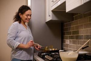 africano americano mujer, contento ama de casa cocineros panqueques en un fritura cacerola, sonrisas dulcemente, en pie cerca el negro estufa en el hogar cocina foto