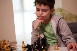 Thoughtful boy of school age keeps his hand on his chin, looks thoughtfully at the chessboard,developing game strategy and moves in a chess game photo