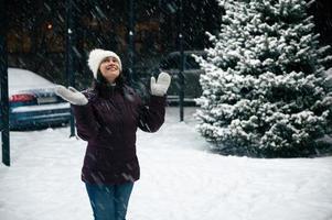 Charming multi-ethnic woman wearing wool mittens and warm clothes, looking up the sky, enjoys the snowfall in the city photo