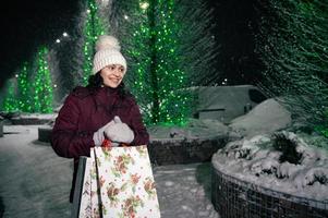 agradable mujer con compras bolsas, caminando abajo el calle, aligerado por festivo iluminación a invierno Nevado noche. foto