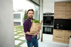 Handsome Caucasian man, charming guy, stands in a home kitchen with a cardboard bag full of healthy food and whole grain bread, looks at camera against the background of windows overlooking the garden photo