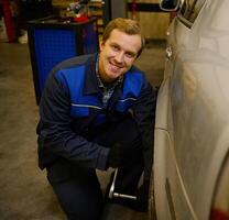 hermoso caucásico hombre, profesional auto mecánico a mano cambios el rueda en el auto, desenroscando el nueces y tornillos con el llave inglesa, sonriente a el cámara en el reparar tienda foto