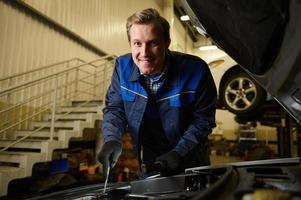 Smiling handsome Caucasian man, professional mechanic, auto technician repairing car engine at car service workshop. photo