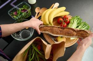 Top view of male hands holding a whole grain freshly baked bread from patisserie and unpacking an eco paper bag with healthy food, putting it on a wooden cutting board for cooking fresh salad photo