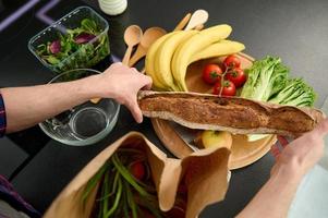 parte superior ver de masculino manos participación un todo grano recién horneado un pan desde pastelería y desempacar un eco papel bolso con sano alimento, poniendo eso en un de madera corte tablero para Cocinando Fresco ensalada foto