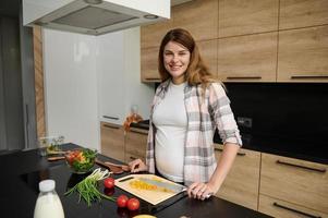 Beautiful Caucasian pregnant woman cutting yellow bell pepper, preparing healthy salad in the home kitchen, cutely smiles looking at camera photo