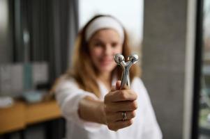 Close-up of a facial massager with two faceted rollers for rejuvenating facial massage in the hands of a blurred red-haired woman in a white bathrobe during her morning routine in the bathroom at home photo