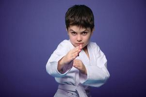 Confident strong kid, aikido fighter practicing martial skills on purple background with advertising copy space. Oriental martial arts practice concept photo