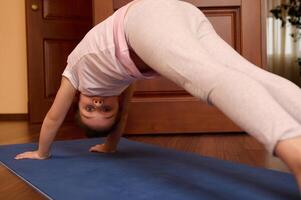 Portrait of 5-6 years old little child girl in downward facing dog pose, looking at camera while practicing yoga at home photo