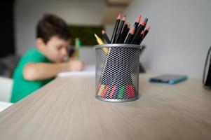 Focus on a pencil holder on a wooden desk against the background of a blurred school boy doing his homework in his room. Education, erudition, smart kids concept photo