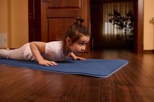 Determined little girl doing push ups, four limbed staff exercise, staying in plank on mat while exrercising indoors photo