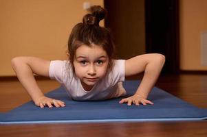 Close-up front view of adorable active little girl doing push ups on a fitness mat at home looking confidently at camera photo