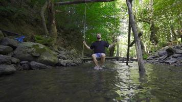 schwingen auf ein schwingen beim das Bach im schleppend Bewegung. Wasser berührt das Füße von das jung Mann schwingen auf das schwingen. video