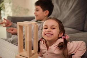 Beautiful child, adorable little girl in pink sweatshirt plays board game with her brother, builds wooden structures from wooden bricks and blocks, smiles toothy smile, looking at camera photo