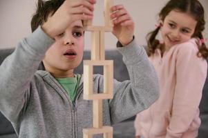 Handsome schoolboy, concentrated on construction of complex tall structure from wooden blocks, against the background of his cheerful little sister, smiling sweetly, looking at his structure. Close-up photo