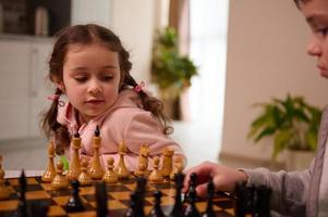 de cerca retrato de un hermosa pequeño europeo niña jugando ajedrez juego con su mayor hermano quien escoge arriba un ajedrez pedazo y hacer su mover. intelectual inteligente desarrollo tablero juegos para niños foto