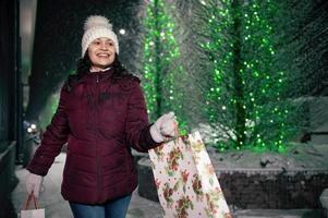 encantador Hispano mujer camina con compras pantalones en un ciudad calle iluminado por guirnaldas a Nevado invierno noche foto