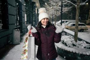 atractivo mujer con compras bolsas, capturas copos de nieve mientras camina a lo largo un Nevado calle iluminado por fiesta guirnaldas foto