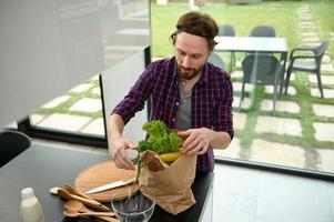 gastos generales ver de un hermoso europeo chico en a cuadros camisa en pie en el cocina en un espacioso villa y desempacar el eco paquete con todo grano junquillo, ensaladas y sano vegetales foto