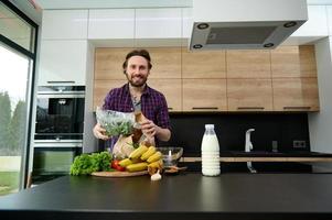 Handsome man smiles looking at camera, holding greens while unpacking eco paper bag with healthy food, standing at kitchen countertop in his spacious villa with large windows overlooking garden photo