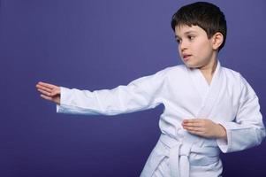 aikido luchador europeo 10 años antiguo chico en blanco kimono mejora su luchando habilidades, aislado en púrpura antecedentes Copiar espacio. oriental marcial letras. foto