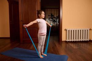 Little child girl in sportswear, standing on a gym mat and doing stretching exercises on arms with elastic band at home photo