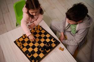 Overhead view of a cute little girl sitting at the table and plays chess with her brother, picking up a chess piece and making his move. Early development, home educational games for children photo