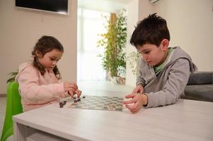 Two adorable Caucasian elementary aged kids, boy and girl, brother and sister having great time playing checkers board game together at home interior. photo