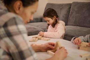 Concentrated little Caucasian girl builds constructions with wooden blocks, developing her fine motor skills, sitting at table with her blurred mother on the foreground, focused on building structures photo