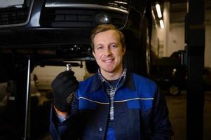 Auto mechanic holding a nut while servicing a car in a workshop, smiles looking at camera against a lifted automobile on a hoist. Car maintenance and auto service concept photo