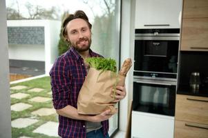 Happy charming Caucasian young man standing in home kitchen with eco paper bag full of healthy food and whole grain bread looking at camera against windows overlooking garden. Healthy eating concept photo