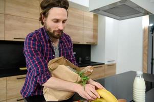Handsome Caucasian man unpacking eco cardboard bag with healthy raw vegan food and a whole grain baguette, bananas and putting ingredients on a kitchen table. Healthy food and lifestyles concept photo