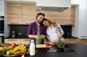 joven embarazada hermosa mujer suavemente pone su cabeza en el espalda de su amoroso marido, preparando para su un sano vegetal ensalada en el hogar cocina. Fresco Fruta mentiras en mesa en primer plano foto