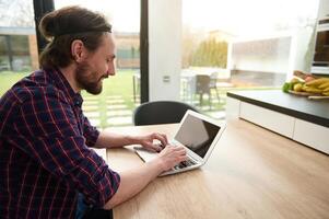 Handsome Caucasian man freelancer in casual attire working remotely on laptop, typing text on laptop with empty blank monitor screen with copy ad space, sitting at table at home during quarantine photo