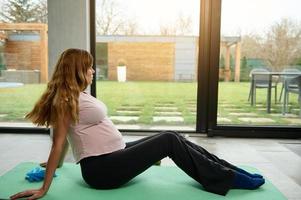 Young pleased active pregnant Caucasian woman exercising at home on a fitness mat. Athletic woman in anticipation of a baby, exercising during pregnancy to maintain a fit and beautiful body photo