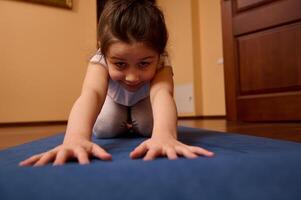 de cerca frente disparo. adorable pequeño niño niña extensión su cuerpo en niño actitud mientras hacer ejercicio en un yoga estera adentro foto