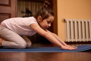 de cerca pequeño niña extensión su cuerpo en balasana pose, sentado en niño actitud mientras practicando yoga en un aptitud estera foto