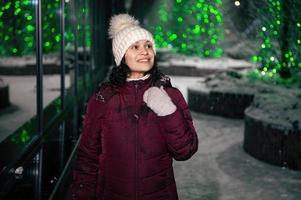 bonito mujer en calentar ropa caminando abajo el calle, iluminado por guirnaldas en un Nevado invierno noche foto