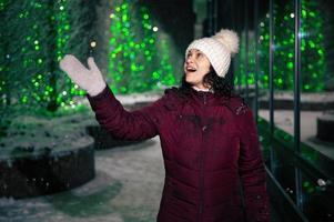 Delightful woman catches snowflakes, enjoys walking down the city street, lightened by garlands on a snowy winter night photo