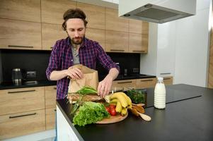 atractivo joven caucásico hombre en casual ropa toma fuera alimento, frutas, verduras, verduras desde un papel eco paquete y pone eso en el cocina mesa, en pie en el cocina de su espacioso casa foto