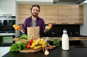 hermoso contento caucásico hombre en casual vestir participación crudo Fresco frutas en manos y sonriente mirando a cámara mientras en pie a cocina encimera detrás eco bolso con comprado sano comida foto