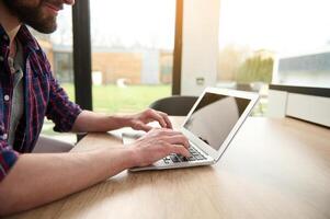 Close-up hands of a man freelancer in casual attire working remotely on laptop, typing text on laptop with empty blank monitor screen with copy ad space, sitting at table at home during quarantine photo